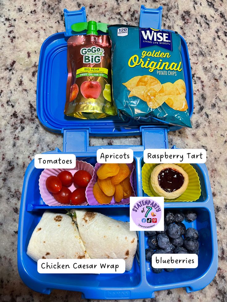 the lunchbox is filled with different foods and snacks, including chips, tomatoes, blueberries, lemons, and tortillas