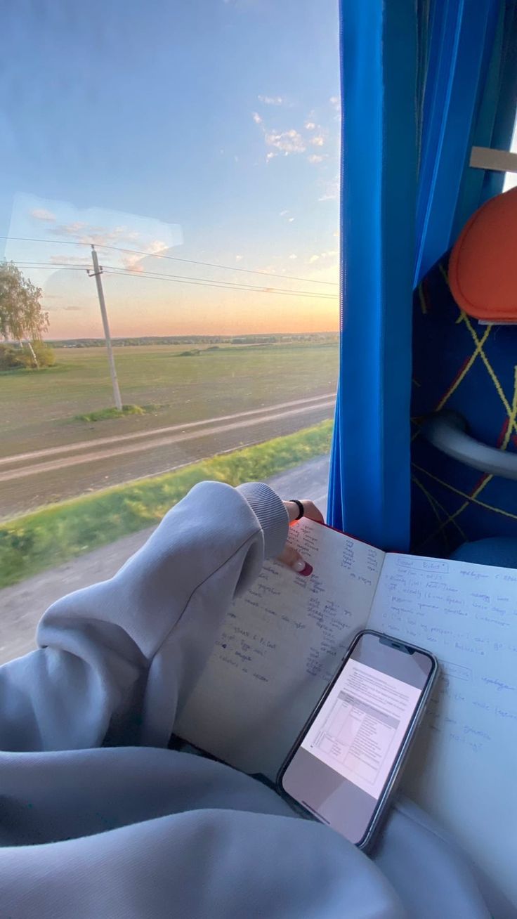 a person sitting on a train looking out the window at an open book and cell phone