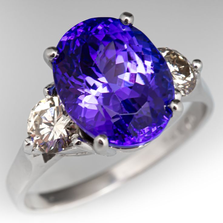 This stunning ring is centered with one (1) oval cut natural tanzanite set into a four-prong setting. The shoulders of the ring are each accented with one (1) prong set round brilliant cut diamond. The ring measures 11.9mm at the top and rises 9.6mm above the finger, tapering to 2.1mm wide and 1.1mm thick at the base of the shank. It currently fits a size 8.75. There is a small scratch on the surface of the tanzanite but it does not detract from the overall magic of the stone. Luxury Tanzanite Ring With Bezel Setting, Luxury Tanzanite Rings With Tension Setting, Luxury Tanzanite Necklaces With Prong Setting, Classic Sapphire Rings In Purple, Oval Tanzanite Gemstones For Formal Occasions, Classic Tanzanite Gemstones For Anniversary, Oval Tanzanite Gemstones In Prong Setting, Brilliant Cut Tanzanite Gemstones, Oval Amethyst Ring With Tanzanite Accent Stones