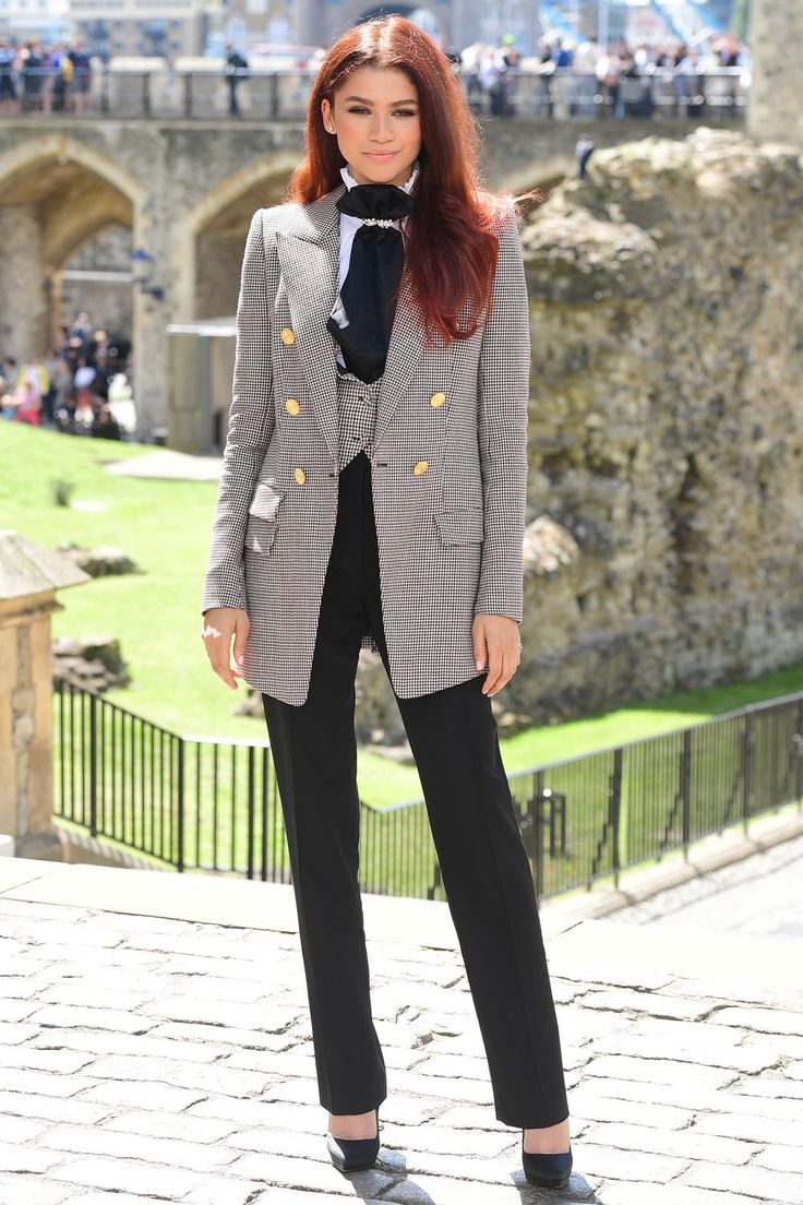 a woman with red hair wearing a gray jacket and black pants standing in front of a castle