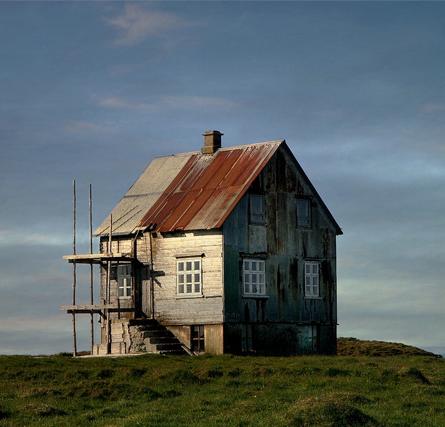 an old run down house sitting on top of a green hill under a cloudy sky