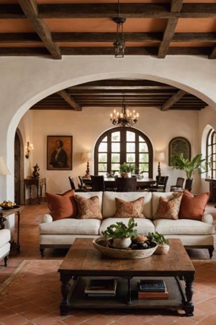 Cozy living room with white sofas and orange pillows, wooden coffee table, arched doorways, and exposed wooden beams on the ceiling. Spanish Style Couch, Mexico Style Living Room, Roman Interior Design Style, Classic Spanish Interior Design, Spanish Villa Interior Living Room, Spanish Style Interior Design Living Room, Spanish Colonial Office, Spanish Hacienda Living Room, Old Spanish Style Homes Living Room