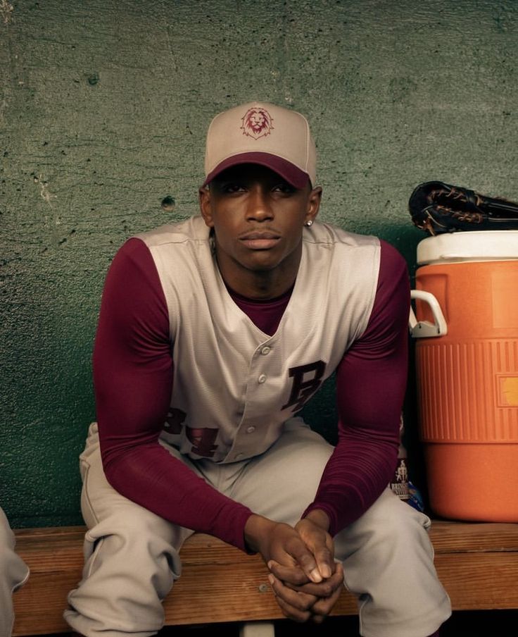 a baseball player sitting on a bench next to an orange cooler and wearing a hat