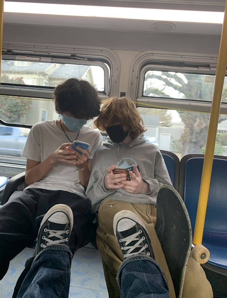 two people sitting on a bus looking at their cell phones while wearing facemasks