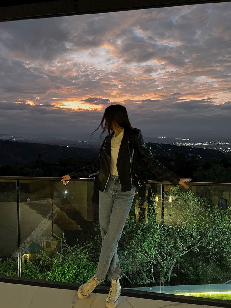 a woman standing on top of a balcony next to a lush green forest under a cloudy sky