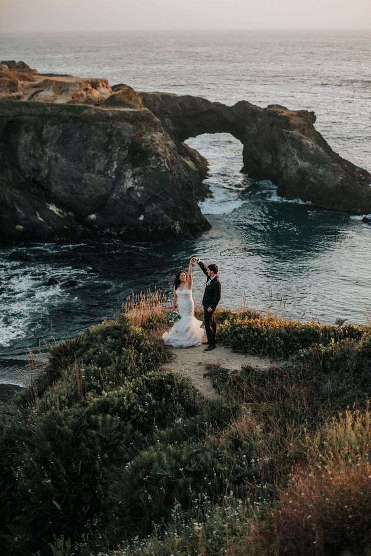 a bride and groom are standing on the edge of a cliff overlooking the ocean with an arch in the background