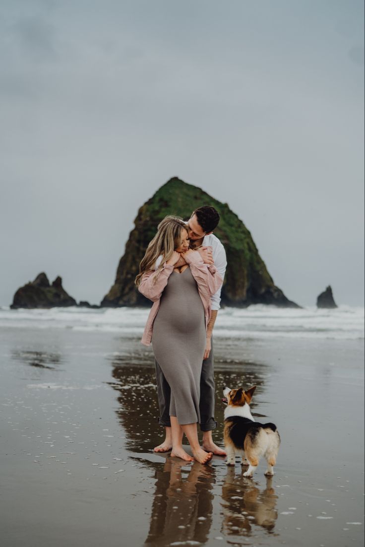 a pregnant woman hugging her husband while standing on the beach with a dog in front of them