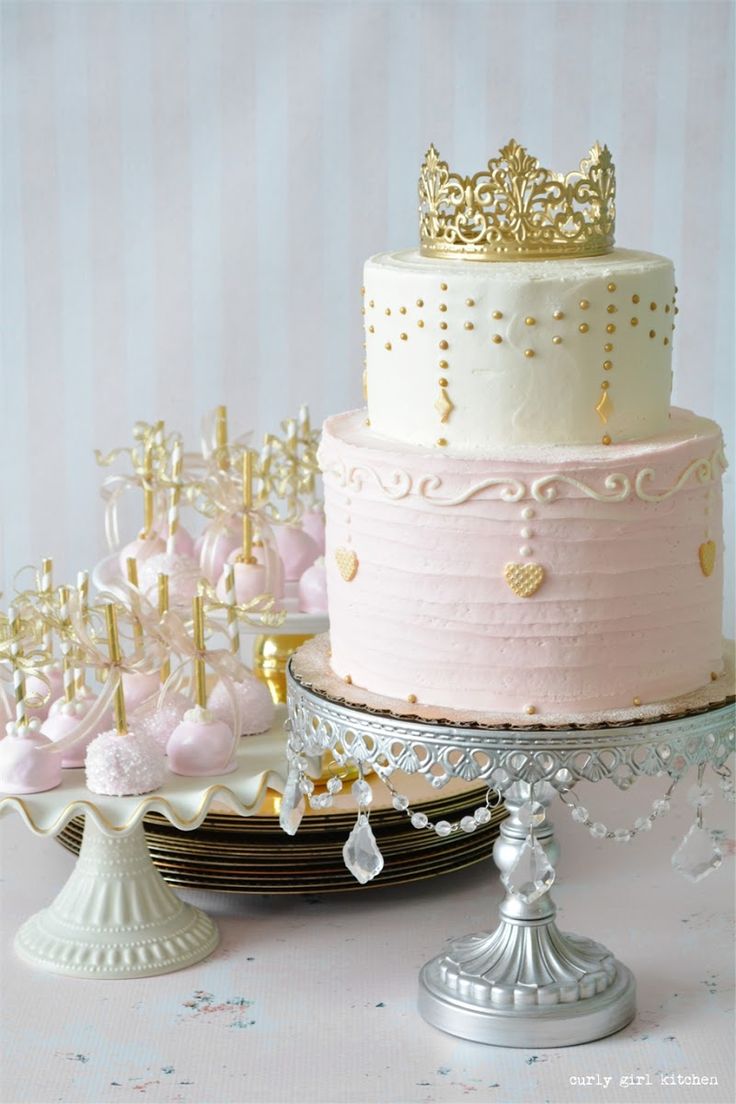 three tiered cake with pink frosting and gold crown on top, surrounded by other desserts