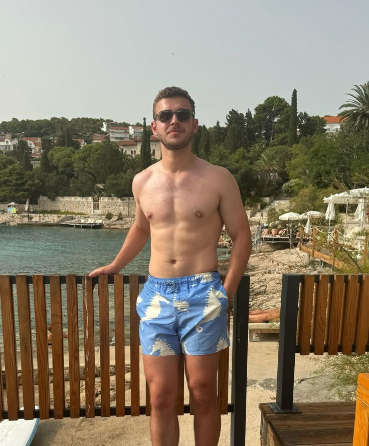 a shirtless man standing next to a wooden fence near the water with his hands on his hips