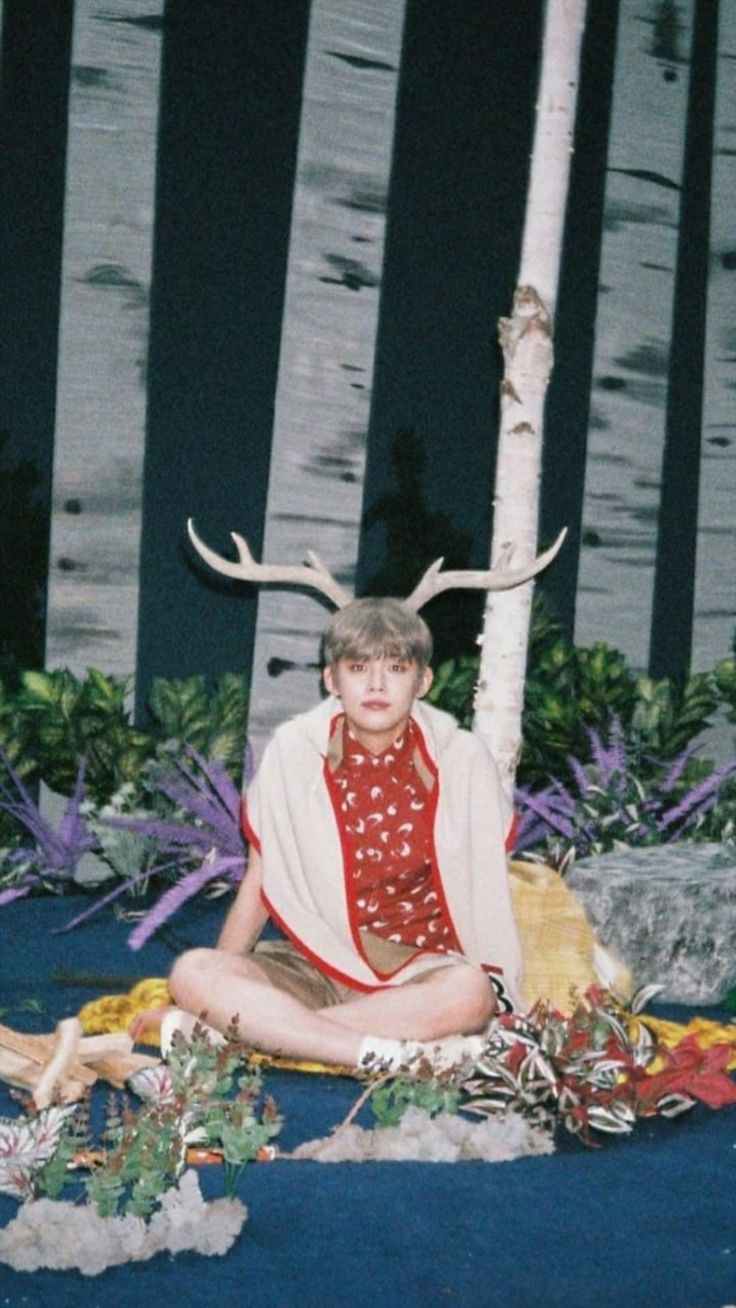 a young man sitting on the ground in front of a tree with antlers around it