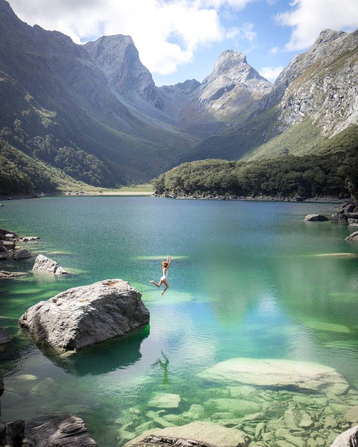 a person jumping into the water from rocks in front of some mountains and green grass