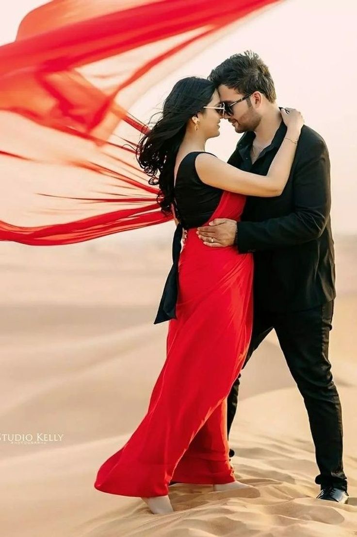 a man and woman embracing in the desert with a red scarf flying above their heads