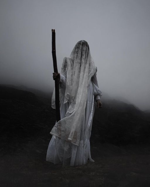 a ghostly woman holding a stick in the fog on a dark mountain side with her face obscured by white veil