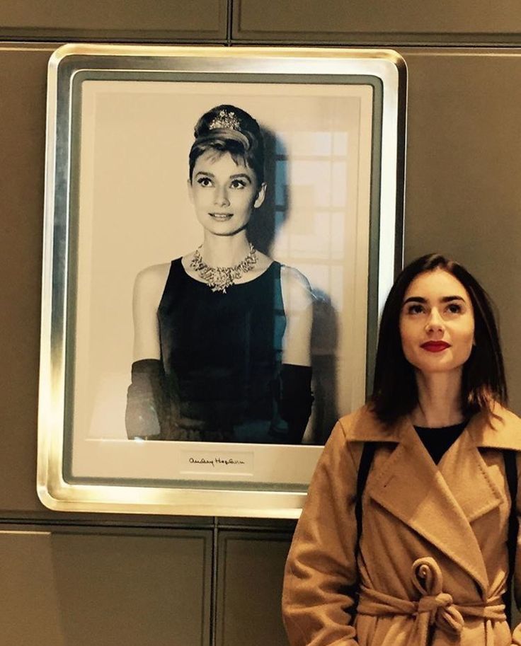 a woman standing in front of a portrait of a woman wearing a black dress and coat