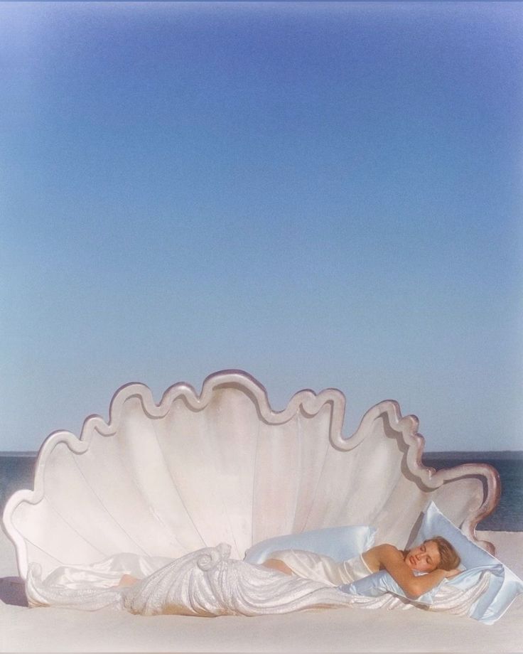 a woman is laying in a large shell on the beach with her head resting on a pillow