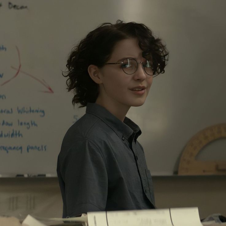 a woman standing in front of a whiteboard wearing glasses and looking at the camera