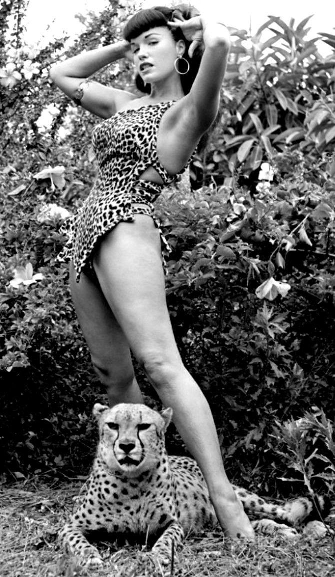 a woman standing next to a cheetah in front of some bushes and trees