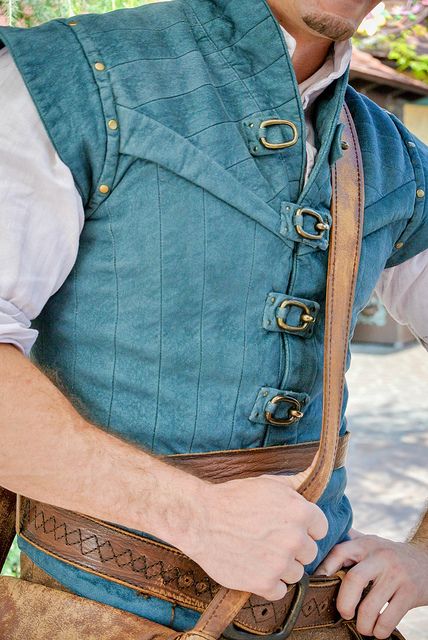 a man wearing a blue vest and hat with his hands in his pockets while standing next to a tree