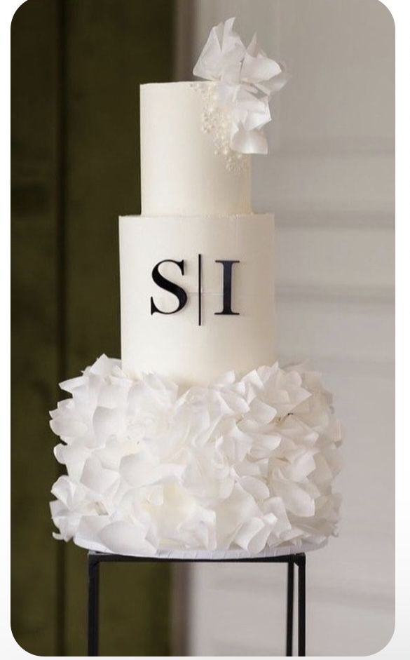 a three tiered white wedding cake with ruffles on the bottom and monogrammed top