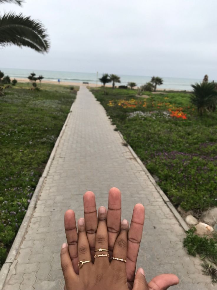 someone is holding their hand up in front of the camera on a path by the beach