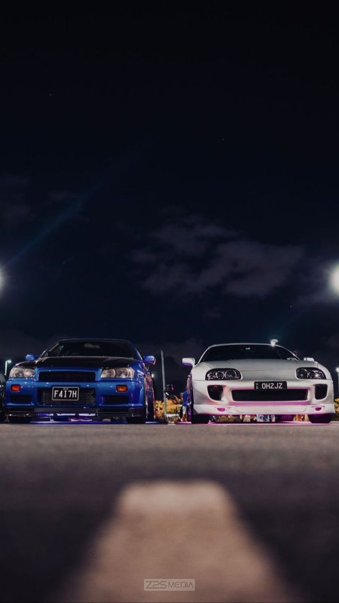 two cars parked next to each other on a street at night with bright lights in the background