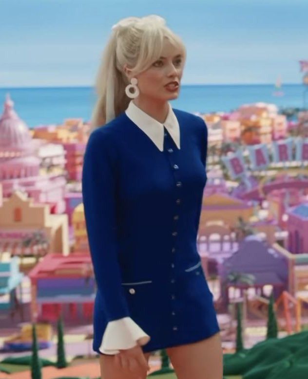 a woman with blonde hair wearing a blue dress and white collared shirt standing in front of a cityscape