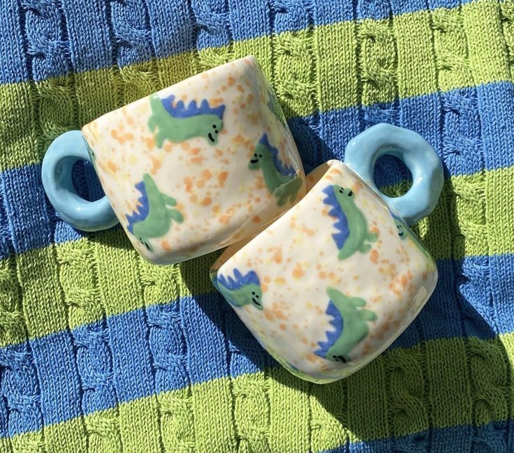 two ceramic mugs sitting on top of a blue and green blanket