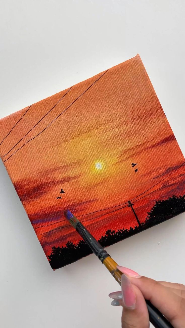 someone is holding a paintbrush in their hand and painting a sunset on the canvas