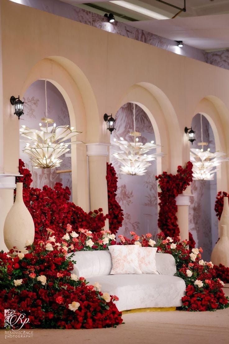 a white couch surrounded by red flowers and chandeliers in a room with arches