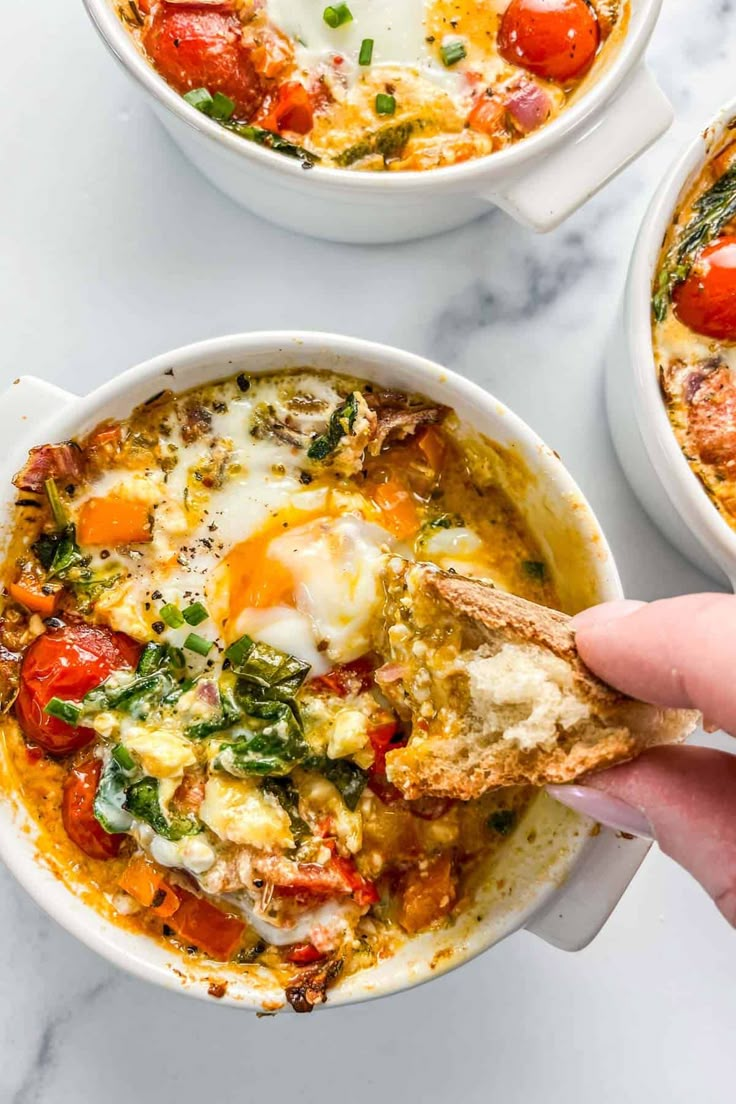 someone holding a piece of bread over a bowl of eggs and tomato soup with spinach