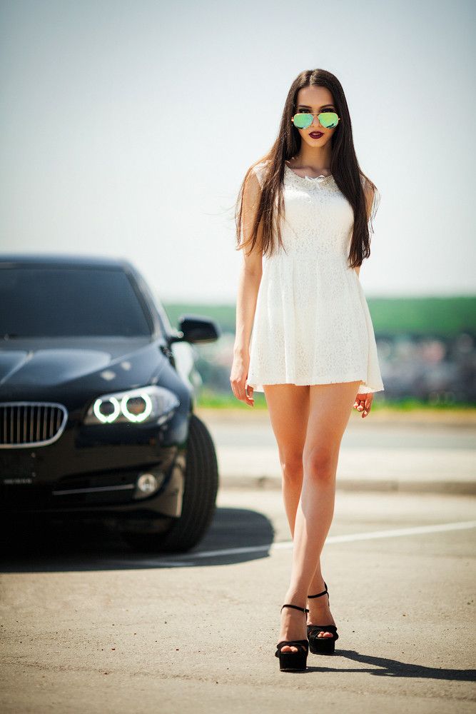 a woman in white dress standing next to a black car with sunglasses on her face