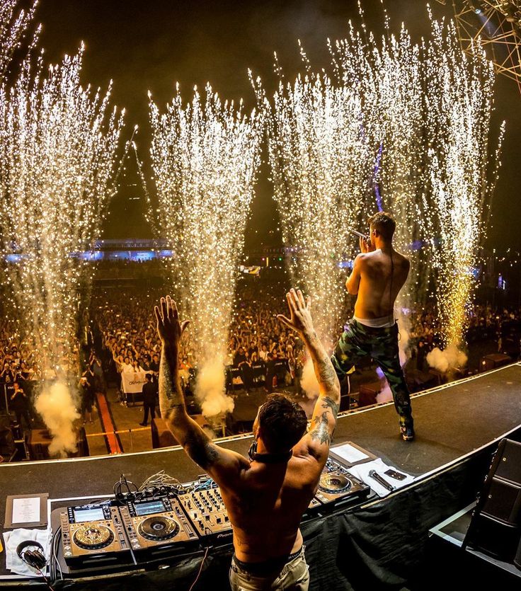 two shirtless men are on stage with fireworks in the sky behind them and dj's holding their hands up