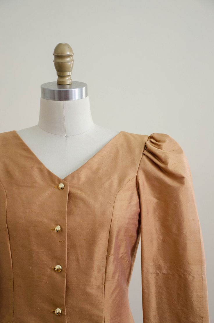 A homemade golden tan jacket, with puffed shoulders, gold faceted buttons, and good tailoring. It is unlined and light enough to be worn as a blouse. ☛ m e a s u r e m e n t s ☚ Bust: 32 Waist: 28 Shoulders: 13.5 Length: 21 ☛ d e t a i l s ☚ Era: 1990s Material: polyester Condition: excellent ☛ v i s i t t h e s h o p ☚ https://etsy.me/2Nd23kg ☛ instagram ┇ poppycockvintage ☛ facebook ┇ poppycockvintage Fitted Tops With Gold Buttons For Workwear, Fitted Gold Top For Workwear, Fitted Gold Tops For Work, Gold Blouse With Buttons For Work, Gold Blouse For Workwear In Fall, Fitted Gold Blouse For Office, Classic Gold Blouse For Workwear, Classic Gold Formal Blouse, Gold Formal Blouse With Buttons