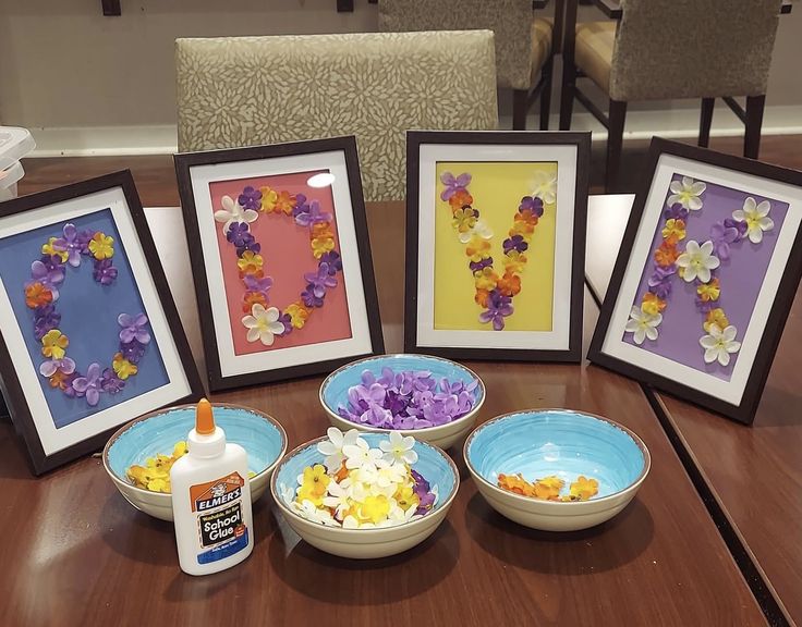 four frames with flowers are sitting on a table next to two bowls and one bottle