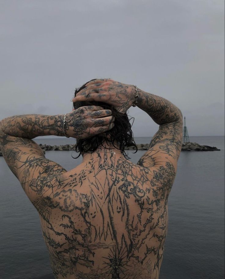 a man with tattoos on his back standing in front of the ocean and holding his hands to his head