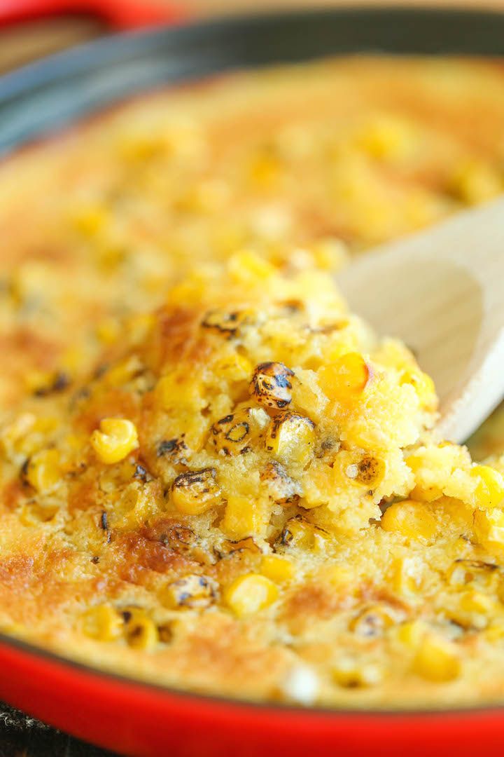 a red bowl filled with corn on top of a wooden table next to a white spoon