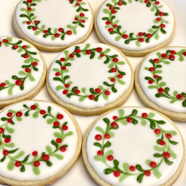 six decorated cookies on a white plate with red berries and green leaves around the edges