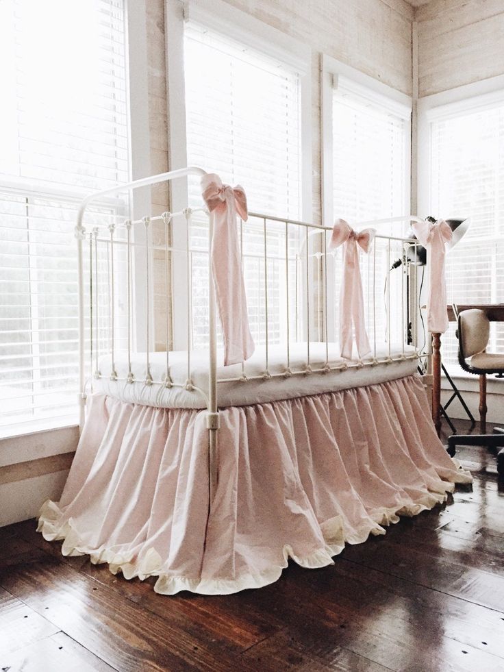 a baby crib with pink ruffles and bows on the bed skirt is in front of two windows