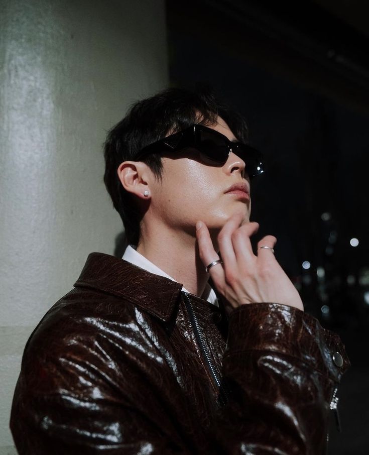 a young man wearing sunglasses and a leather jacket looks off to the side with his hand on his chin