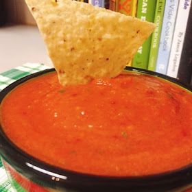 a tortilla chip being dipped into a bowl of tomato soup with a bookcase in the background