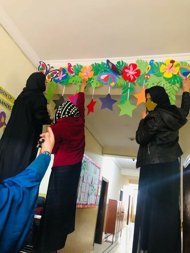 three women in hijabs are hanging decorations from the ceiling with stars and flowers on them