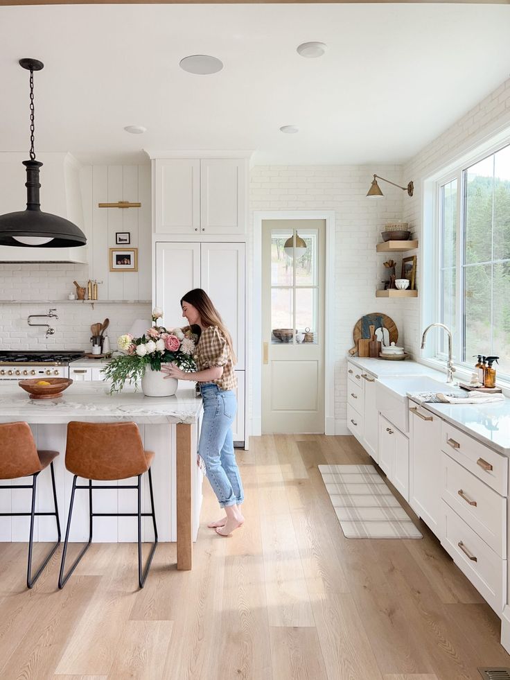 a woman standing in the middle of a kitchen with white cabinets and counter tops, surrounded by brown bar stools