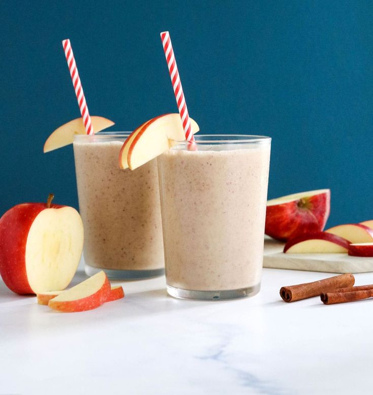 two glasses filled with apple smoothie next to sliced apples and cinnamon sticks on a table