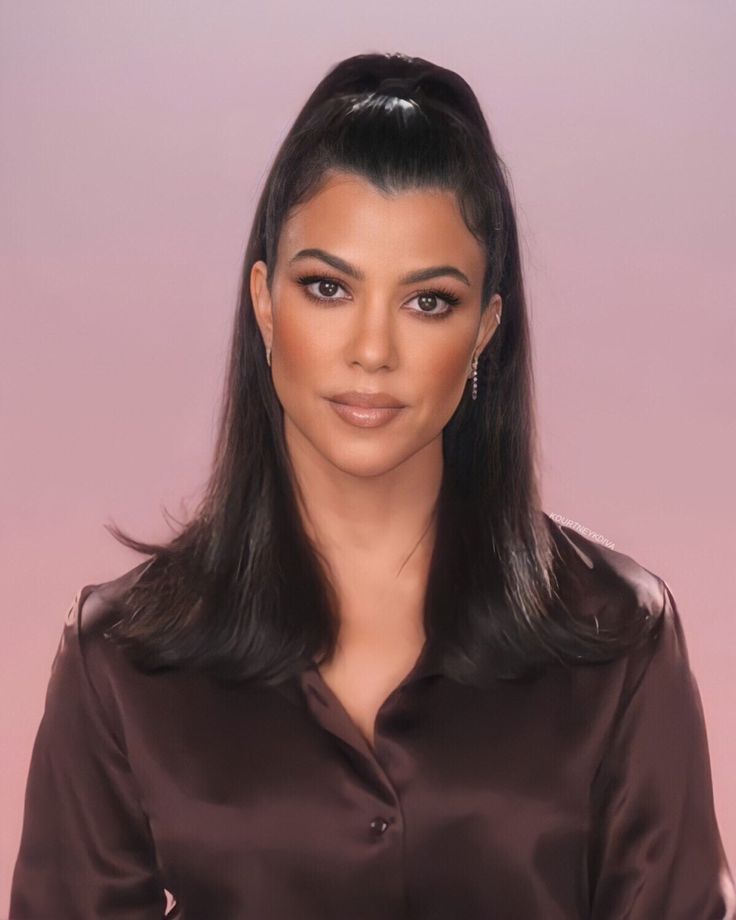 a woman with long black hair wearing a brown shirt and diamond earrings, standing in front of a pink background