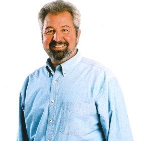 a man in a blue shirt and tie standing with his hands in his pockets, smiling