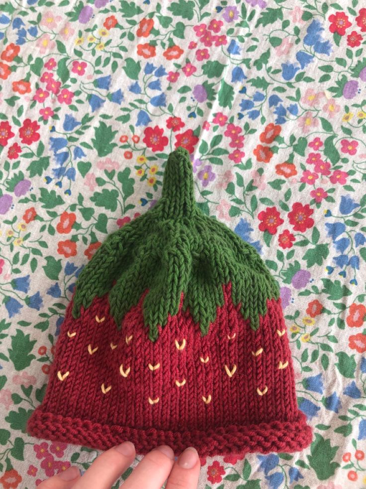 a knitted strawberry hat sitting on top of a floral cloth covered tablecloth with a person's hand holding it