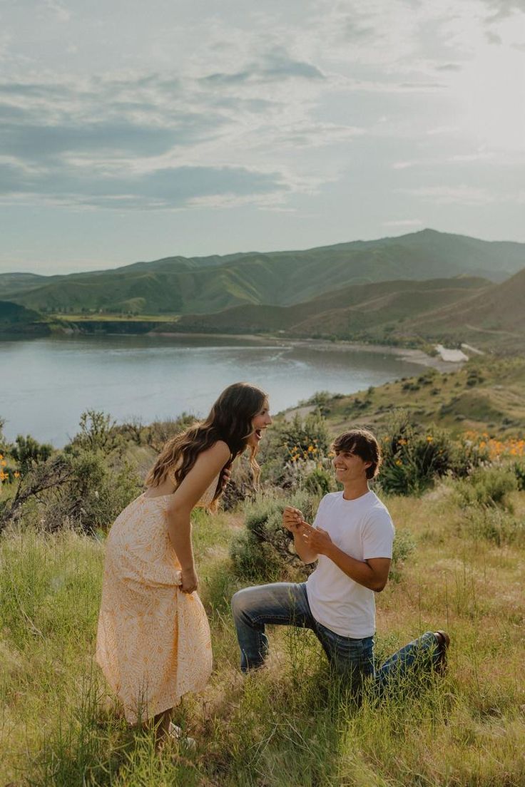 a man kneeling down next to a woman on top of a grass covered hillside near a lake