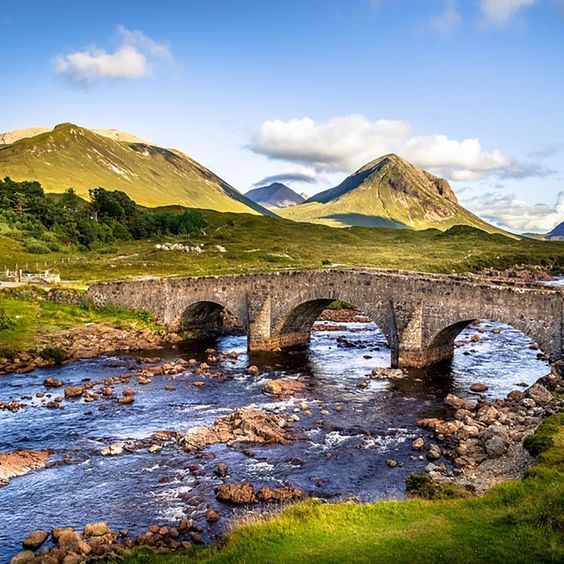 an old stone bridge over a river in the middle of mountains with green grass and rocks