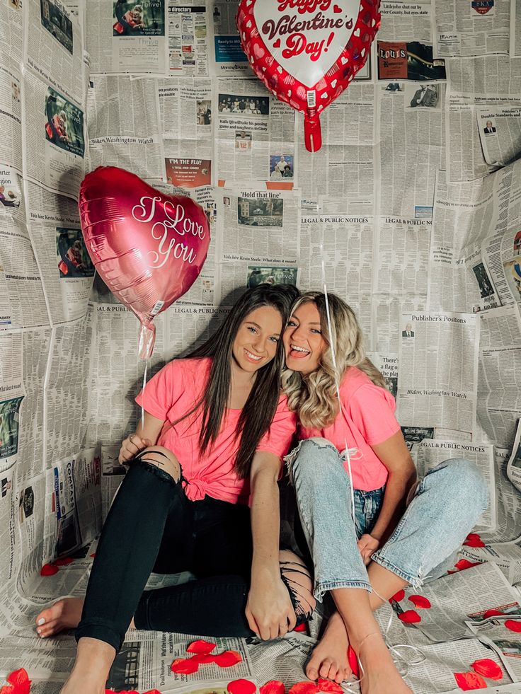 two women sitting on a bed with red balloons and paper hearts hanging from the wall