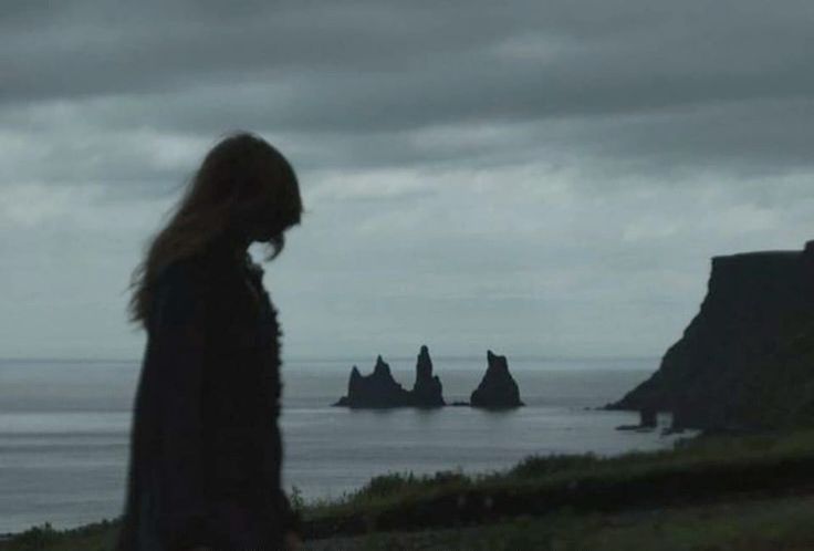 a woman standing on top of a lush green hillside next to the ocean under a cloudy sky